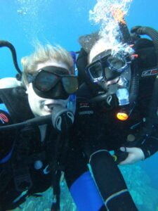 Chelsey and Anthony scuba diving underwater, wearing full dive gear and smiling at the camera.