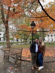 Chelsey and Anthony standing in a park during autumn, surrounded by trees with colorful fall leaves