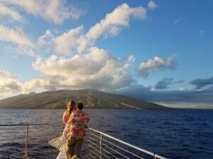 Chelsey and Anthony standing on the deck of a catamaran during sunset, with the island of Maui and wind turbines in the background