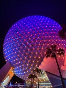 Epcot's Spaceship Earth illuminated with colorful lights at night, with palm trees in the foreground.