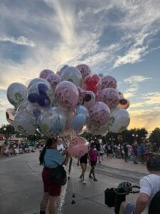 Disney cast member holding a large cluster of Disney-themed balloons at sunset in Magic Kingdom, with Anthony riding an ECV in the foreground.